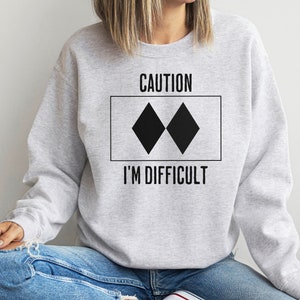 May include: A grey sweatshirt with the text "CAUTION" and a black double diamond symbol. The text "I'M DIFFICULT" is below the symbol.