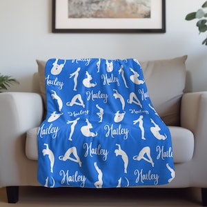 May include: A blue blanket with white yoga poses and the name "Hailey" in a cursive font. The blanket is draped over a beige armchair. The background includes plants and a framed picture.