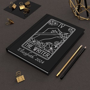 May include: Black journal with a white illustration of a hand holding a pen and flowers. The text "THE WRITER" and "Elizabeth 2024" are printed in white on the front cover.