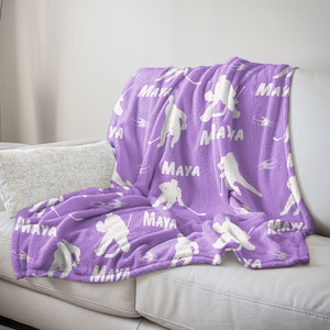 Peut inclure: Une couverture en polaire violette avec un motif de silhouette de joueur de hockey blanc et le nom "Maya" répété partout.