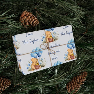 Puede incluir: Papel de regalo blanco con un tema de Winnie the Pooh. El papel presenta globos azules, un tarro de miel y un oso sosteniendo un tarro de miel. El texto en el papel dice "Love, The Taylors" y "Congratulations Ashley and Sean!"