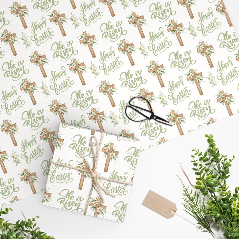 May include: White wrapping paper with a repeating pattern of a wooden cross with white flowers and the text "He is Risen" and "Happy Easter".