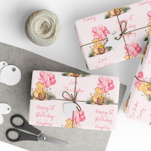 May include: Pink and white wrapping paper with a Winnie the Pooh character holding a jar of honey and pink balloons. The text "Happy 1st Birthday Kingsley" is printed on the paper.  The paper is tied with a brown string and a white tag.