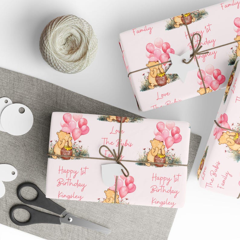 May include: Pink and white wrapping paper with a Winnie the Pooh character holding a jar of honey and pink balloons. The text "Happy 1st Birthday Kingsley" is printed on the paper.  The paper is tied with a brown string and a white tag.