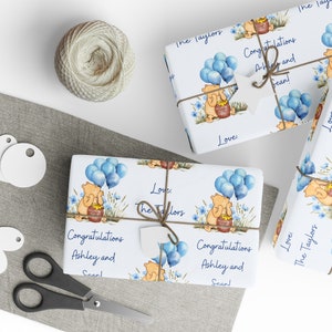 Puede incluir: Un papel de regalo blanco con un dise&ntilde;o de globos azules y blancos. El papel presenta un personaje de dibujos animados de Winnie the Pooh sosteniendo una olla de miel. El texto en el papel dice "&iexcl;Felicidades Ashley y Sean!" y "Con amor: Los Taylors".