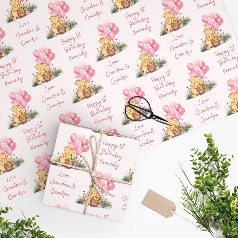 May include: Pink and white wrapping paper with a Winnie the Pooh design. The paper features a cartoon Winnie the Pooh holding a pot of honey with pink balloons. The text on the paper reads "Happy 1st Birthday Kennedy" and "Love Grandma & Grandpa".