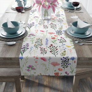 May include: A white table runner adorned with a watercolour floral design, showcasing diverse wildflowers in pink, purple, red, and yellow hues. The runner is placed on a wooden table, complemented by place settings, including plates, bowls, and wine glasses.
