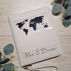 Könnte beinhalten: Cremefarbenes Reise-Portemonnaie mit einem schwarzen Weltkarten-Design. Die Geldbörse hat eine Handschlaufe und einen goldfarbenen Reißverschluss. Die Namen "Marc & Bianca" sind auf der Vorderseite aufgedruckt.