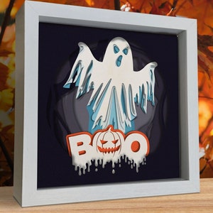 May include: A white framed picture with a 3D paper cut-out design of a ghost and the word "BOO" with a pumpkin face in the center. The background is dark blue.