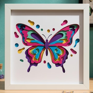 May include: A framed, multi-layered paper art piece featuring a colorful butterfly design. The butterfly has layers of pink, purple, yellow, and blue, with cut-out details and a white frame. The artwork is on a wooden surface.