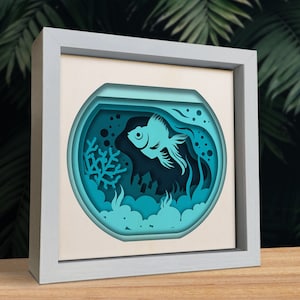 Op de afbeelding: Een blauwe papieren uitsnijding van een vis in een kom met koraal en bubbels. De vis is in 3D-stijl en lijkt te zwemmen.