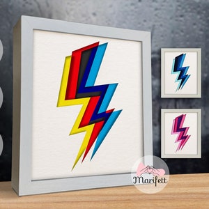 Może przedstawiać: Wydruk w ramie przedstawiający kolorowy wzór błyskawicy. Błyskawicę tworzy warstwowy papier w odcieniach żółtego, czerwonego, niebieskiego i czarnego. Wydruk znajduje się w białej ramie z białą passe-partout.