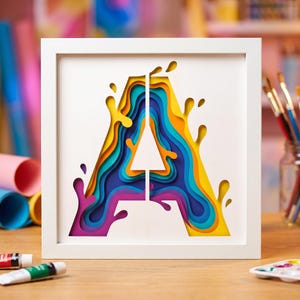 May include: A square white frame containing a layered paper art piece of the letter A. The design features a multi-colored gradient effect with shades of yellow, orange, blue, purple, and teal, creating a depth effect with liquid-like splashes.