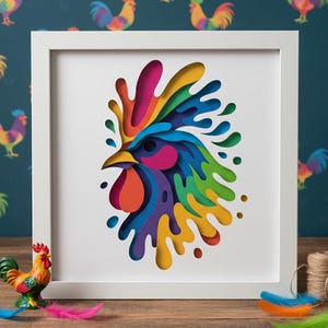 May include: A framed art print featuring a colorful rooster design. The rooster is depicted in a layered paper cut style, with vibrant hues of blue, yellow, orange, pink, and purple. The artwork is set against a white background within a white frame.