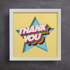 May include: A framed artwork featuring the words "THANK YOU" in a layered, colourful design. The text is set against a light blue star shape, with a yellow background, within a white frame.