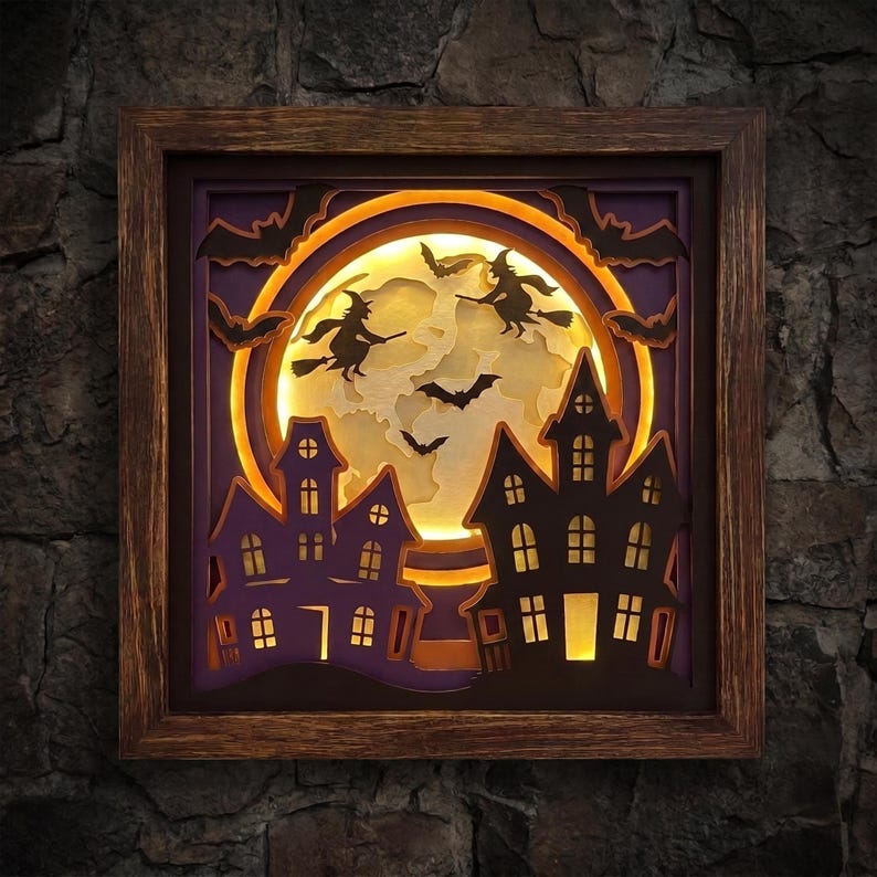 May include: A square wooden framed shadow box featuring a layered paper cutout scene of two silhouettes of witches on broomsticks and several bats flying in front of a large, illuminated yellow moon behind two dark purple haunted houses.