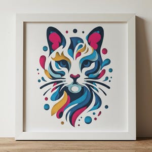 Op de afbeelding: Een ingelijste kunstdruk met een gestileerde kattenkop. Het kunstwerk maakt gebruik van gelaagde papieren uitsparingen in blauw, roze, geel en wit. De ogen van de kat zijn blauw en het witte frame versterkt de levendige kleuren.