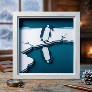 May include: A framed paper art piece featuring a penguin standing on an iceberg, with its reflection in the water below. The artwork is in a white frame and has a layered design with shades of blue and white. A compass and pen are on the wooden surface.