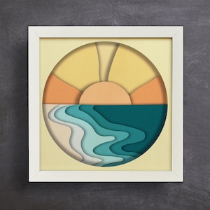 May include: A framed paper art print with a layered design of a sunset over a blue ocean. The sun is yellow and orange, and the ocean is a deep blue with white waves.