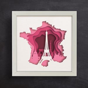 May include: A white paper cutout of the Eiffel Tower in Paris, France, with a pink and white paper cutout of the outline of France behind it. The Eiffel Tower is the focal point of the image, with the outline of France serving as a backdrop.