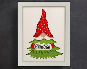 May include: A white frame with a red and white gnome holding a sign that says "Christmas" in black lettering. The gnome is wearing a red hat with white polka dots and has a white beard. The gnome is standing in front of a green Christmas tree.