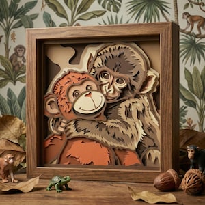 May include: A layered paper art piece in a wooden frame depicts two hugging monkeys. The artwork uses shades of brown and orange, creating a 3D effect. The frame sits on a wooden surface with decorative leaves and small figurines.