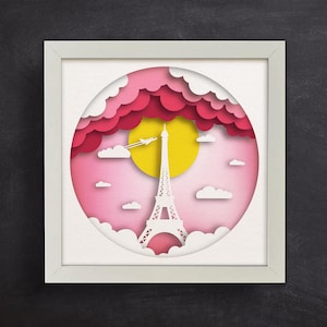 May include: A white frame with a pink and yellow paper cut-out design of the Eiffel Tower in Paris, France. The design includes clouds and a plane flying overhead.
