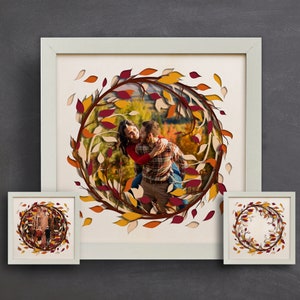 Op de afbeelding: Een wit frame met een uitgesneden ontwerp van herfstbladeren in tinten rood, oranje en geel. De bladeren vormen een cirkel rond een foto van een stel in een herfstdecor.