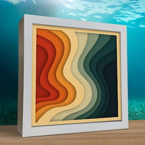 May include: A framed paper art piece with a retro colour palette. The artwork features wavy, contoured layers in shades of red, orange, yellow, green, and blue, encased in a white wooden frame. The piece is displayed on a wooden surface.