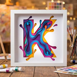 Könnte beinhalten: Ein quadratischer weißer Rahmen mit einem mehrschichtigen Papierkunstwerk des Buchstabens K. Das Design zeigt fließende, überlappende Formen in leuchtenden Farben wie Violett, Blau, Pink, Gelb und Orange, die einen dreidimensionalen Effekt erzeugen.
