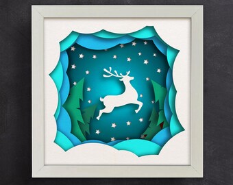 May include: A white paper reindeer leaps through a blue and green paper cut-out winter scene. The scene is framed in a white frame.