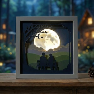 Può includere: Un'opera d'arte in carta incorniciata che raffigura una coppia seduta su una panchina sotto un albero, illuminata da una luna luminosa. L'opera presenta una cornice grigia, con un paesaggio blu scuro e verde. La luna è di un giallo brillante e caldo.