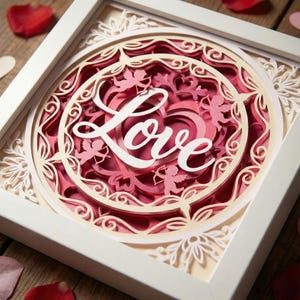 May include: A framed paper art piece with a layered design. The artwork features the word "Love" in white script, set against pink and cream layers. Decorative swirls and cherub figures add detail to the romantic theme.