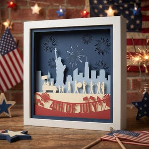 Peut inclure: Une œuvre d'art encadrée sur papier à thème patriotique. L'œuvre présente un design en couches avec un paysage urbain, des feux d'artifice et le texte "4th of July". La palette de couleurs comprend le rouge, le blanc et le bleu, avec un cadre blanc.