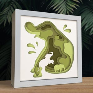 May include: A framed paper art piece depicting elephants in a layered green landscape. The artwork uses varying shades of green to create depth, with a white elephant silhouette in the foreground. The frame is a light grey colour.