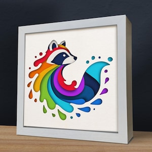 Op de afbeelding: Een wit ingelijst wandkunstwerk met een kleurrijk papiergesneden ontwerp van een wasbeer met een regenboogstaart. De wasbeer kijkt naar links en de staart is om de kop van de wasbeer gewikkeld.