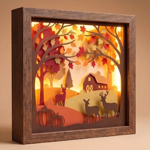 May include: A layered paper shadow box with a dark wood frame featuring an illuminated autumn scene. The display shows silhouettes of deer, pumpkins, trees with fall foliage, and a barn, all backlit to create a warm, glowing layered effect.