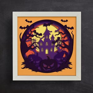 May include: A framed Halloween-themed paper cut art piece. The artwork features a spooky purple haunted house, bats, trees, and a jack-o'-lantern against an orange and yellow background. The artwork is framed in a white square frame.