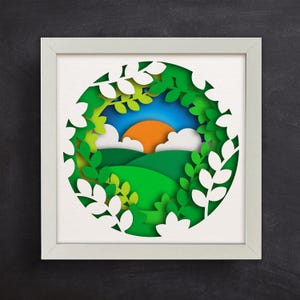 Op de afbeelding: Een wit frame met een ontwerp van een uitgestanste papieren groene landschap met een blauwe lucht, witte wolken en een oranje zon. Het landschap is omringd door witte papieren bladeren.
