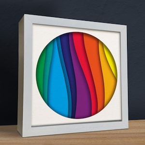May include: A white framed picture featuring a colourful, layered paper design in the shape of a circle. The circle is made up of different shades of green, blue, purple, red, orange, and yellow.
