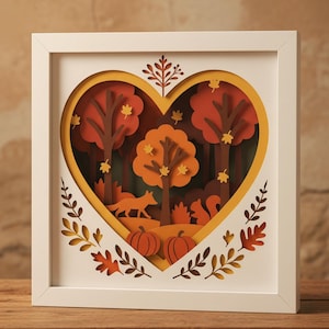May include: A framed paper art piece depicting an autumnal forest scene within a heart shape. The artwork uses layered paper cutouts to create trees, a fox, a squirrel, pumpkins, and falling leaves in orange, brown, and yellow, set in a white frame.