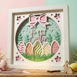 May include: A white framed Easter-themed paper art piece. The design features a pink bow, the words "Happy Easter", and several decorative Easter eggs in a circular design. The background is light green with floral and leaf cutouts.