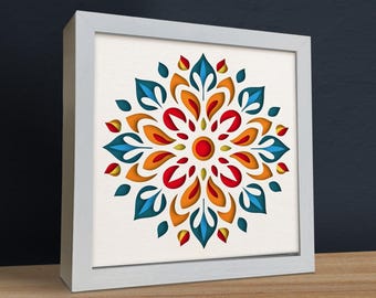 May include: A framed, colorful mandala art piece. The design features layered, symmetrical shapes in shades of red, orange, yellow, and blue. The artwork is set against a white background and framed in a white square frame.