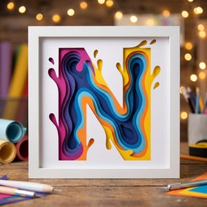 May include: A square white frame containing a multi-layered paper art piece of the letter N. The design features a depth effect with concentric cutouts in vibrant shades of pink, purple, orange, yellow, and various tones of blue, creating a liquid splash effect.