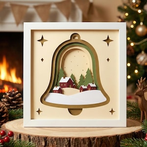 May include: A framed paper art piece featuring a bell shape with a winter village scene. The artwork includes red houses, green trees, and a snowy landscape. The frame is white, and the background is a light beige color.