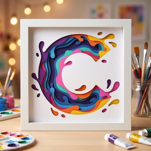 May include: A square white frame containing a multi-layered paper art piece featuring a large letter C. The design uses stacked cutouts in vibrant shades of purple, blue, teal, pink, orange, and yellow, creating a dimensional splash effect.