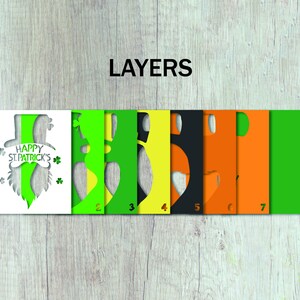 May include: Seven layers of paper cutouts for a St. Patrick's Day card. The top layer is white with the text "Happy St. Patrick's" and a green leprechaun hat and beard. The remaining layers are green, yellow, black, orange, and green.