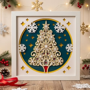 Könnte beinhalten: Ein gerahmtes Weihnachtsbild. Das Kunstwerk zeigt ein mehrschichtiges Design mit einem weißen Rahmen. Der Baum ist gold und weiß, vor einem tealfarbenen Hintergrund mit Schneeflocken-Akzenten. Der Rahmen hat gelbe Diamant-Ausschnitte.