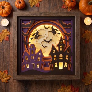 May include: A square wooden shadow box frame containing a layered paper cutout scene. The design features a large glowing yellow moon, black silhouettes of two witches on broomsticks, flying bats, and two dark purple haunted houses with lit windows.