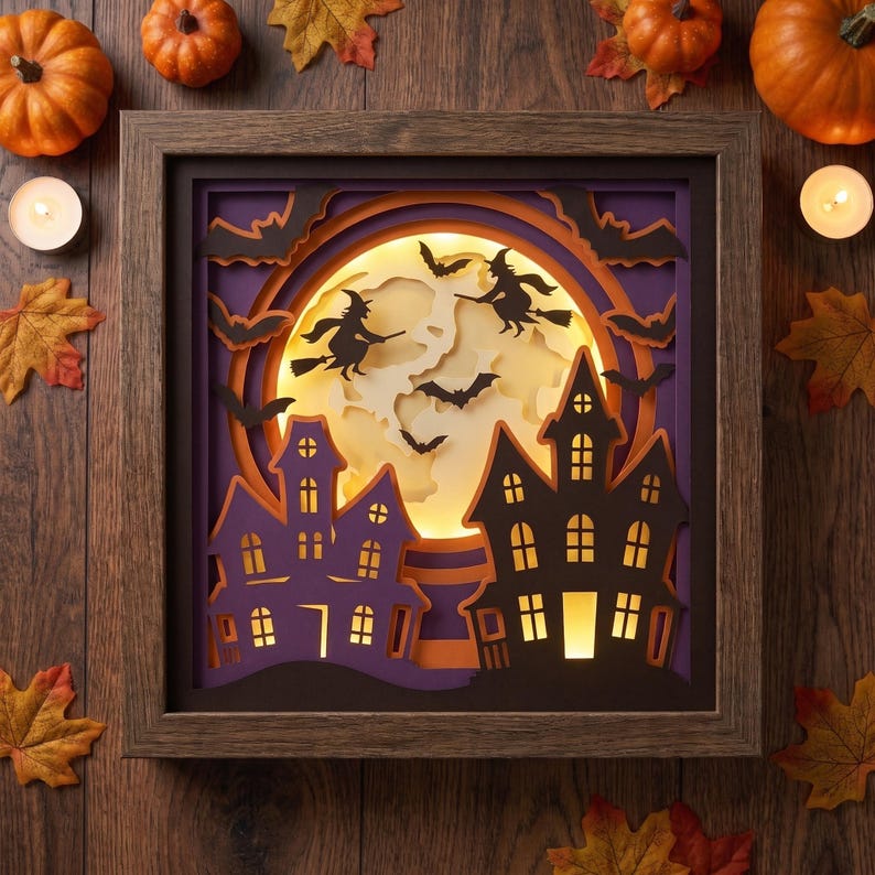 May include: A square wooden shadow box frame containing a layered paper cutout scene. The design features a large glowing yellow moon, black silhouettes of two witches on broomsticks, flying bats, and two dark purple haunted houses with lit windows.